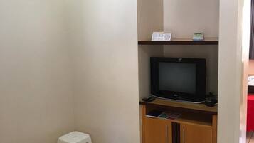A room with a television on a wooden stand, a white stool, and a cabinet with a red table in the background.