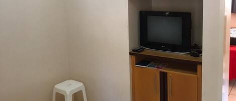 A room with a television on a wooden stand, a white stool, and a cabinet with a red table in the background.