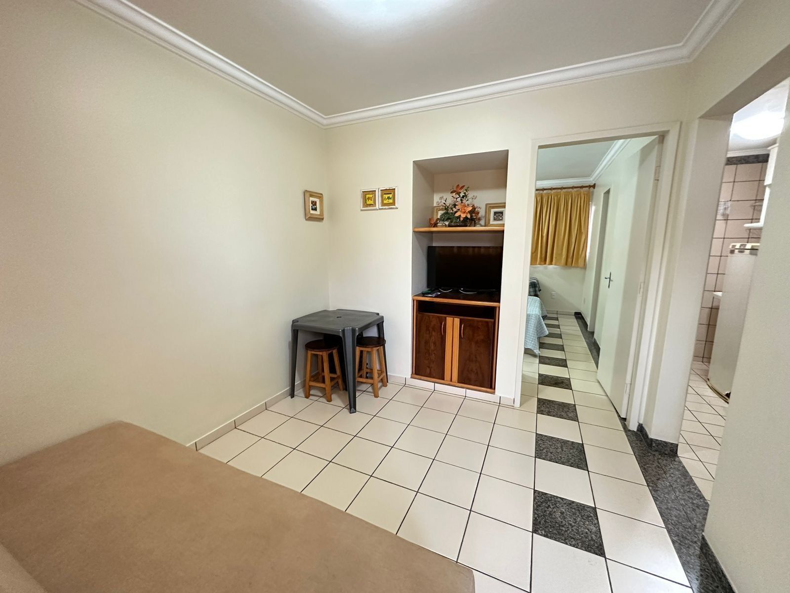 Una habitación pequeña y limpia, con piso de baldosas, una mesa de madera, dos taburetes, una kitchenette pequeña y una puerta que da a otra habitación.