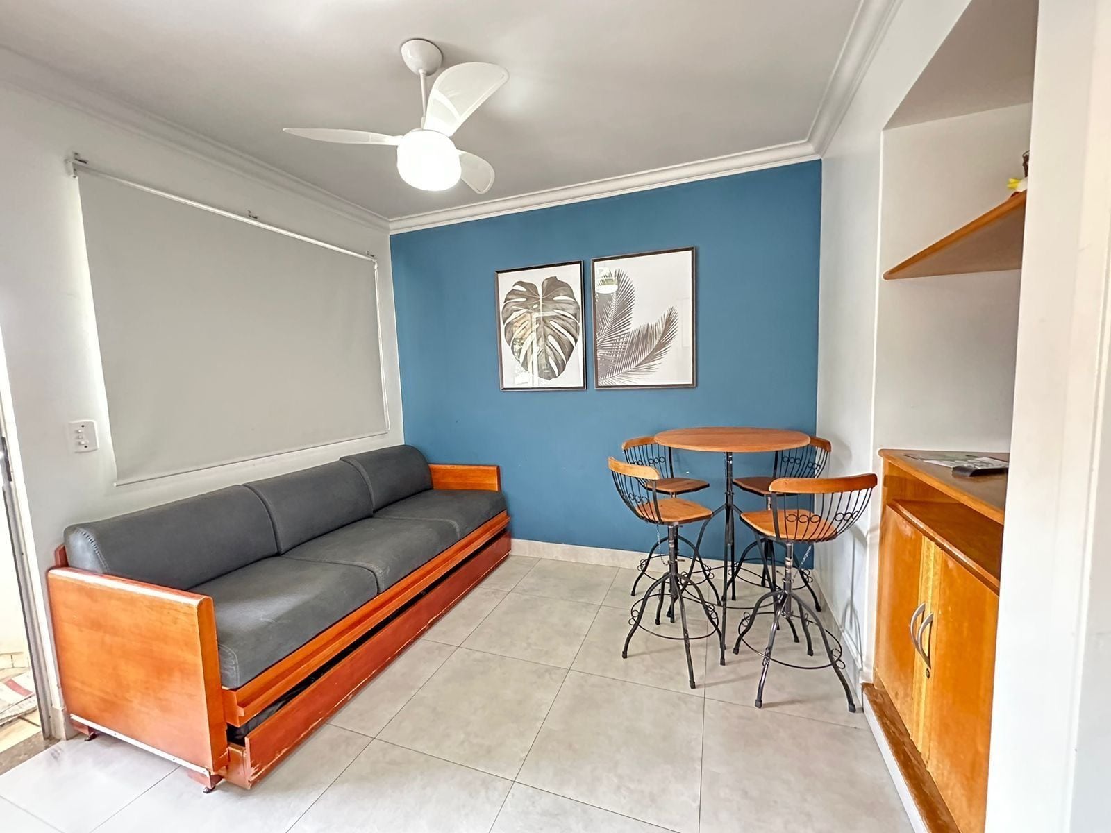A room with a grey sofa, a ceiling fan, two framed leaf artworks, a small round table with chairs, and a wooden cabinet.