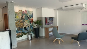 A modern reception area with a large abstract mural, a reception desk, a seating area with a blue chair, and an air conditioning unit on the wall. - DiRoma International Resort Via Caldas (Caldas Novas)