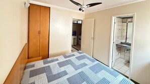 Une chambre à coucher avec un lit, une armoire, un ventilateur de plafond et une salle de bain visible à travers une porte ouverte.