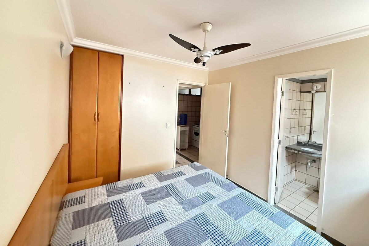 Ein Schlafzimmer mit Bett, Kleiderschrank, Deckenventilator und einem durchgehenden Türbogen einsehbaren Badezimmer.