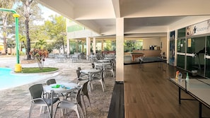 Uma área de pátio externa com mesas, cadeiras, piscina e mesa de pingue-pongue.