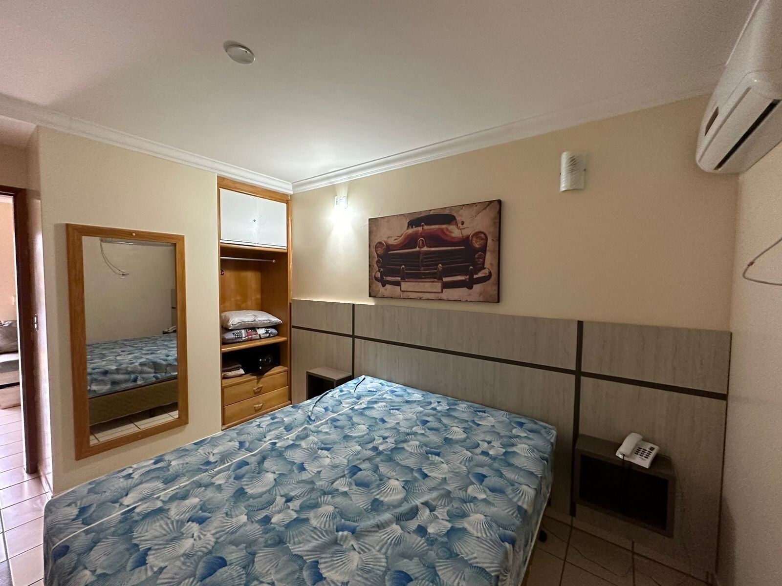 Een slaapkamer met een bed, een kledingkast, een spiegel, een schilderij en een telefoon op een plank.