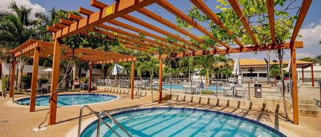 A pool area with wooden pergolas, lounge chairs, and a clear blue pool.