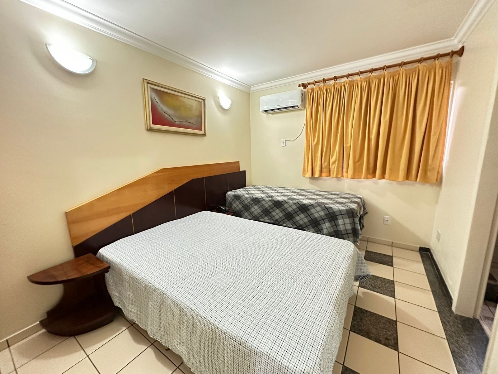 Een slaapkamer met een bed, nachtkastje, een wandairconditioner, een raam met gordijnen en een schilderij aan de muur.