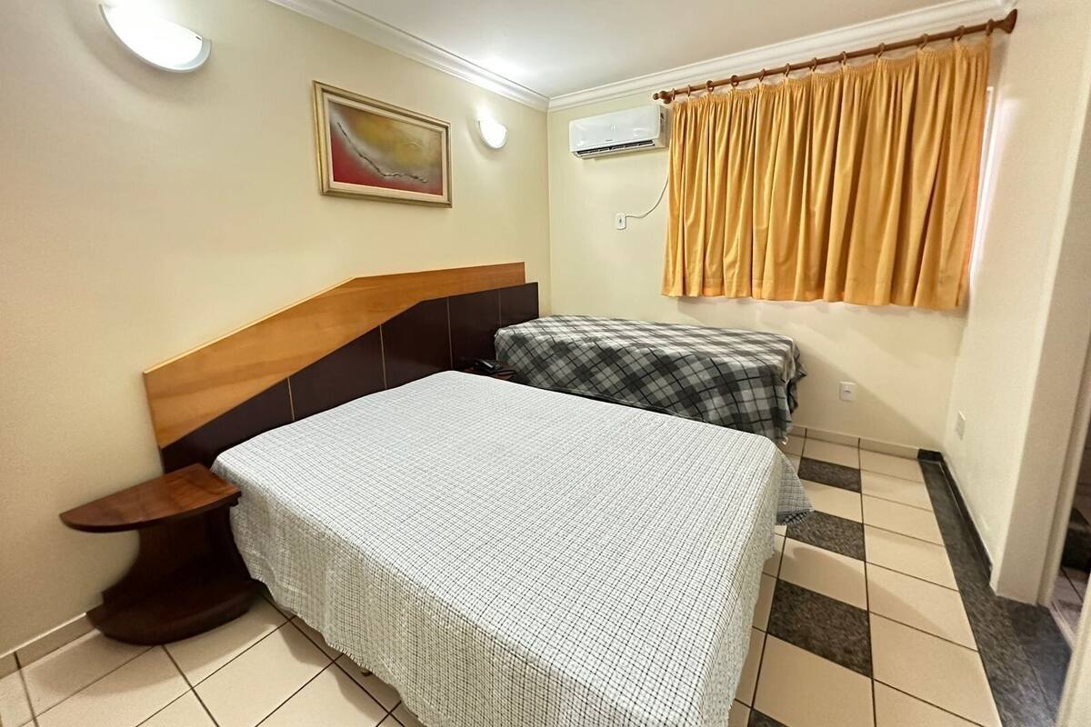 Quarto com cama, mesa de cabeceira, ar condicionado fixado na parede, janela com cortinas e um quadro na parede.