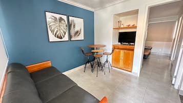A living room with a sofa, a small dining table with chairs, a TV, and artwork on the wall.