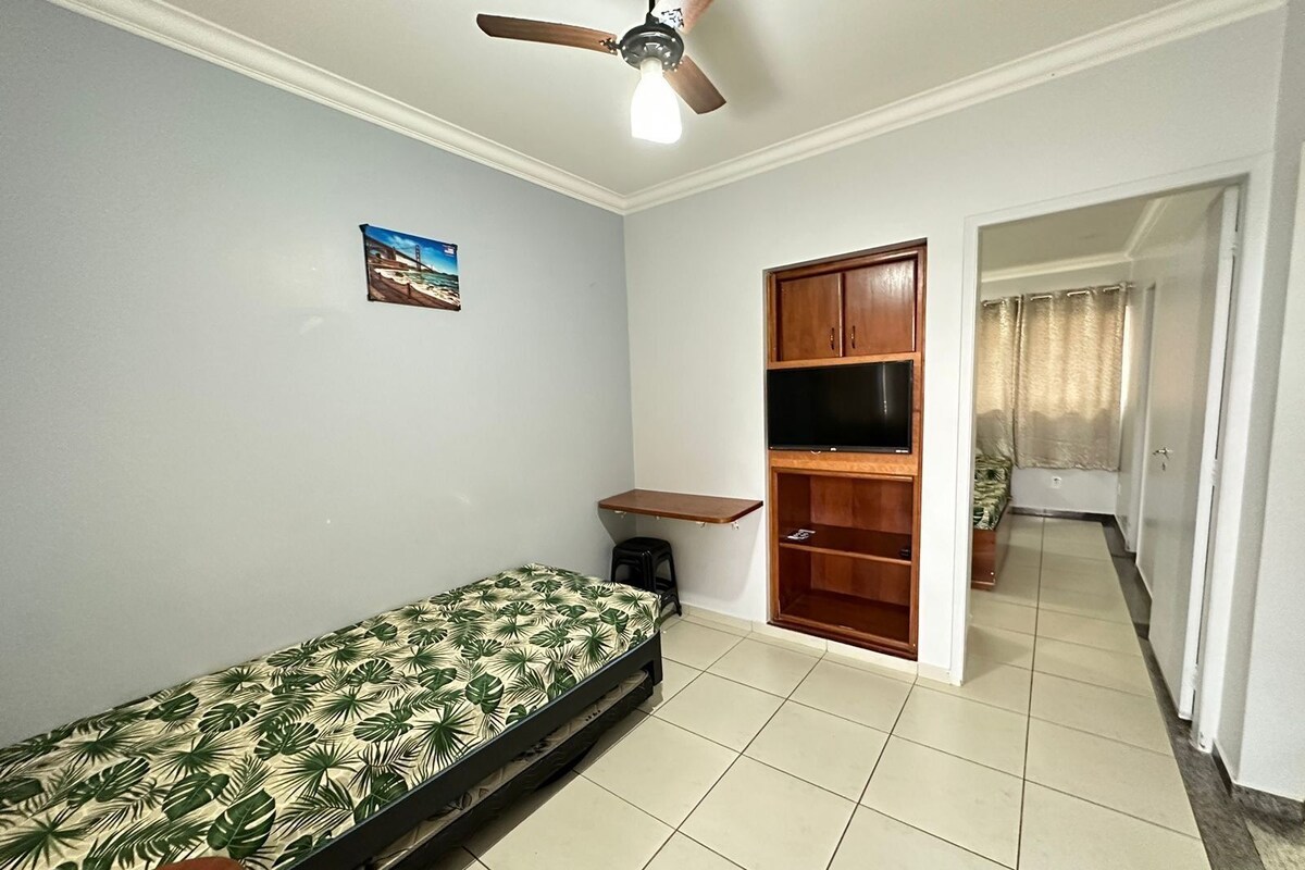 Ein Zimmer mit einem Bett, einem Deckenventilator, einem Fernseher auf einem Holzregal und einem kleinen Schreibtisch.