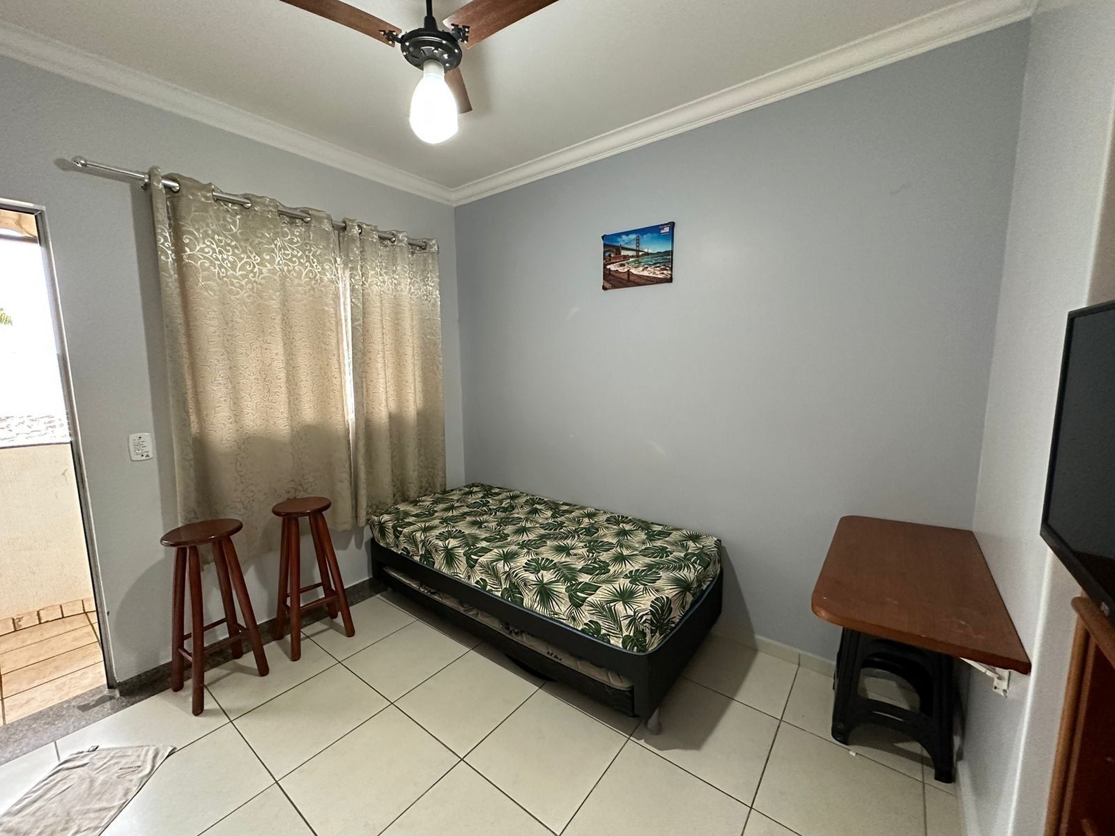 Une petite pièce comprenant un lit, deux tabourets, un ventilateur de plafond, un téléviseur et un tableau accroché au mur.