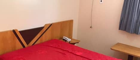 A bedroom with a bed, a bedside table, a telephone, a wall-mounted air conditioner, and a window with curtains.