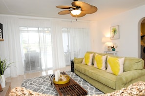 A living room with a green sofa, a ceiling fan, a coffee table with decorative items, and a view through large windows. - The Island House of Siesta (Sarasota)