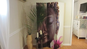 Ett rum med en vit vägg, ett trägolv, en vit byrå med lådor, en tv som är monterad på väggen och en stor dekorativ panel med en bild av Buddha och en rosa blomma.