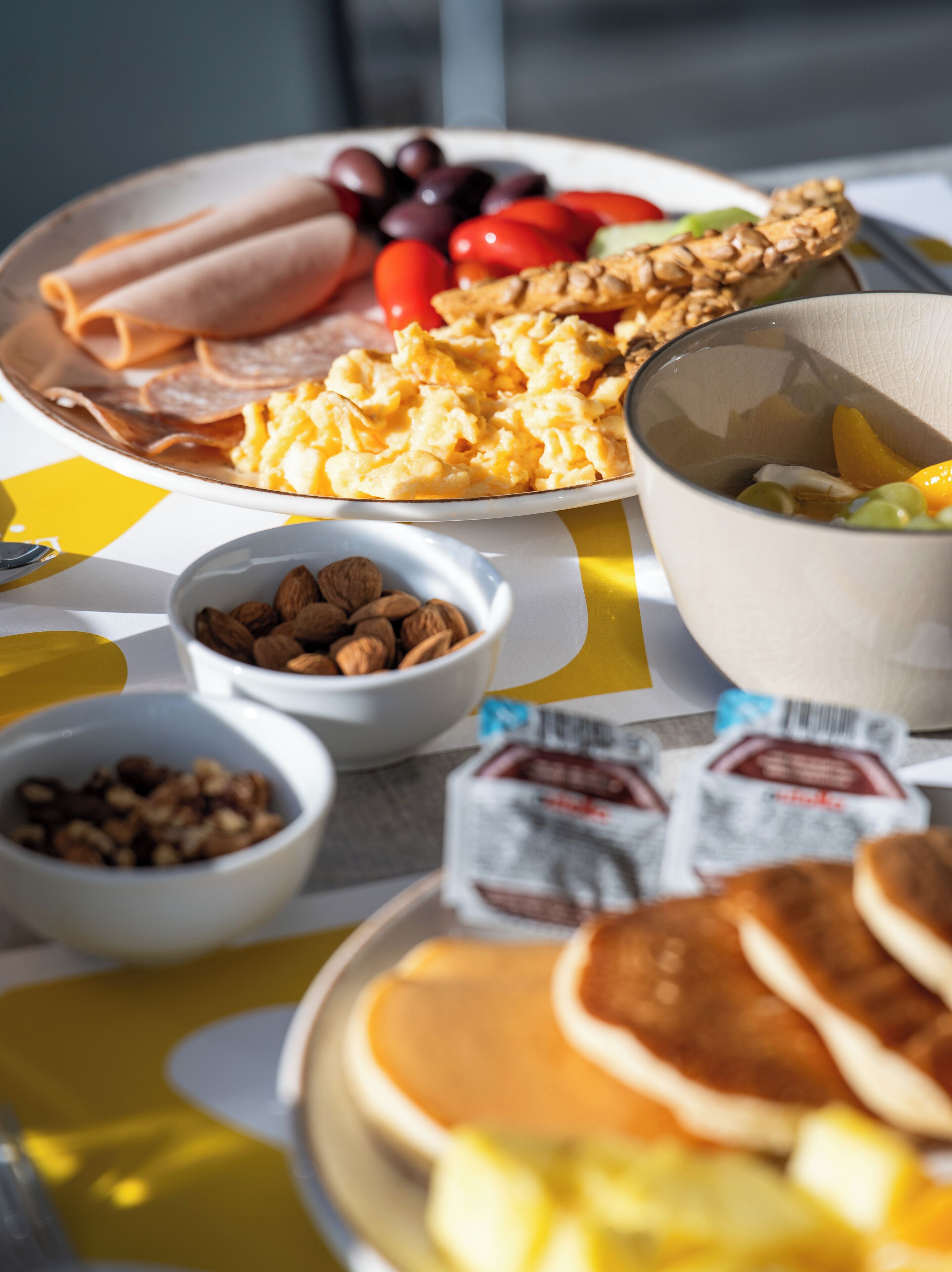 A breakfast table with scrambled eggs, bacon, tomatoes, and a bowl of cereal.