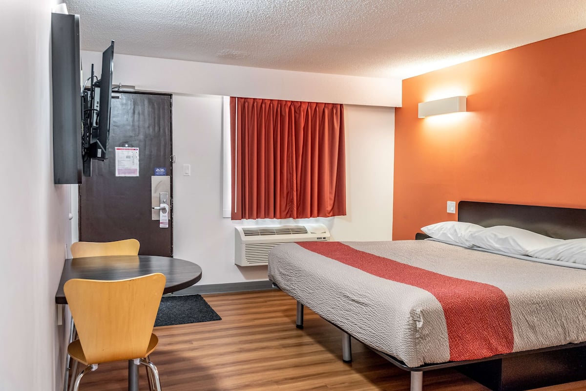 A hotel room with a bed, a desk, a chair, a TV mounted on the wall, and a window with curtains.