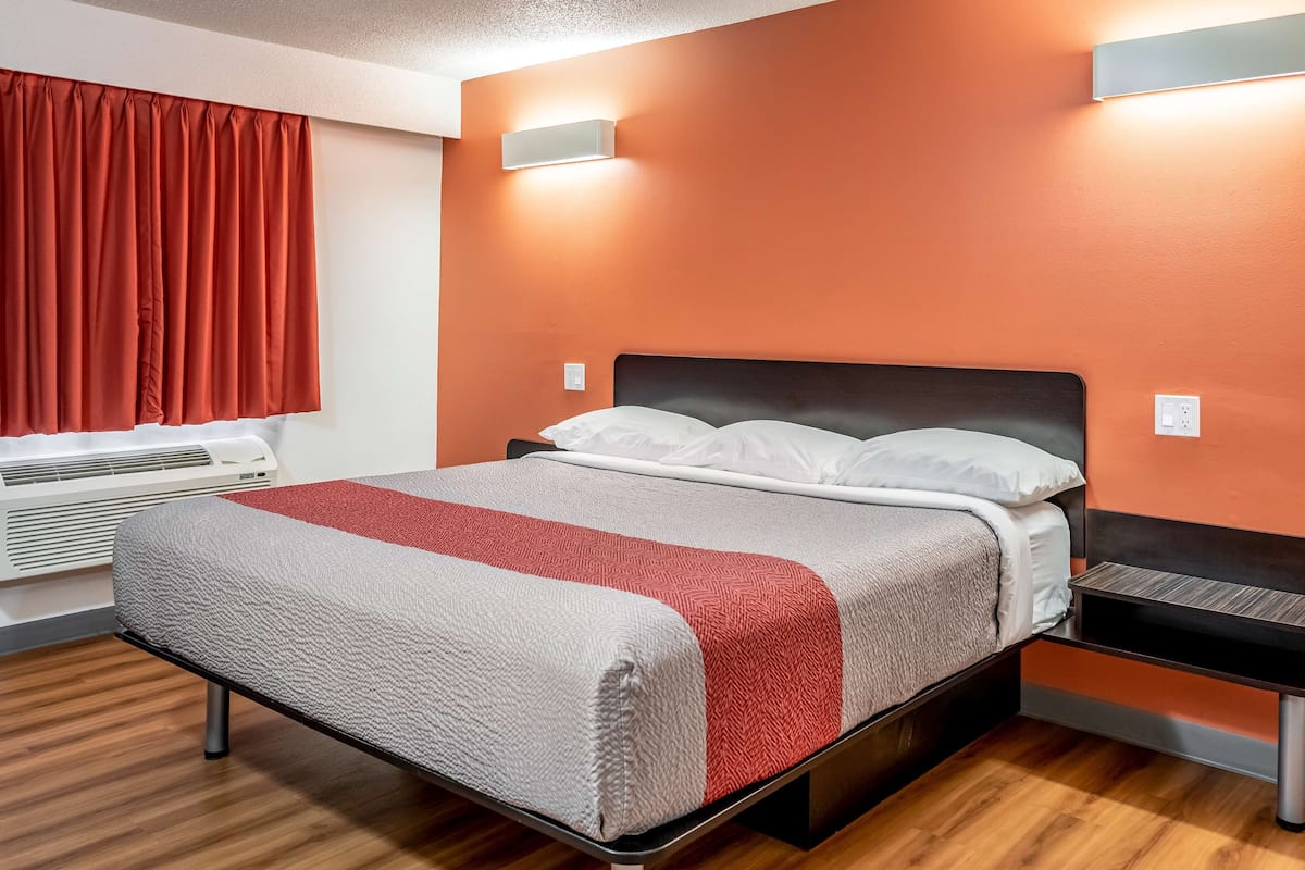 A modern hotel room with a bed, a red curtain, an air conditioning unit, and a wall-mounted light fixture.