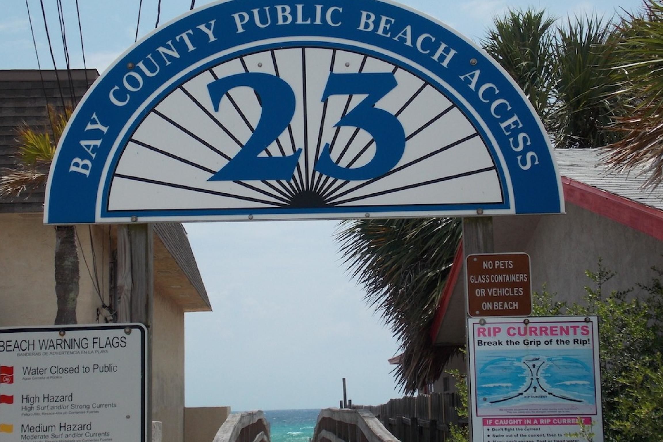 A beach entrance sign with the number 23, a warning flag sign, and a rip current safety poster.