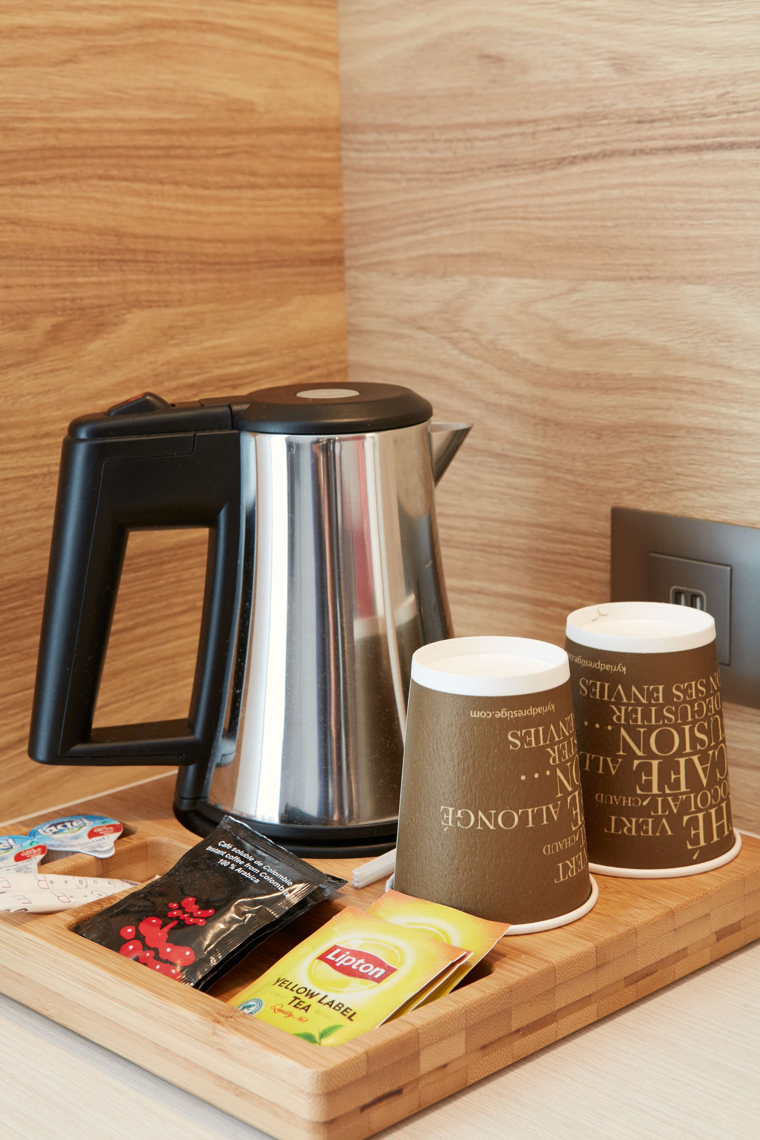 A stainless steel kettle, two disposable cups, a packet of instant coffee, and a Lipton tea packet on a wooden tray.