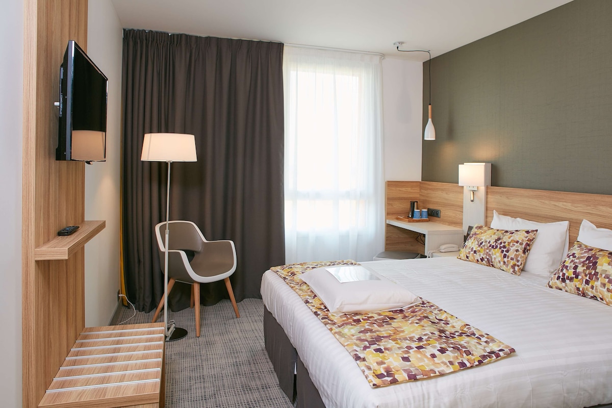 A hotel room with a bed, a desk, a chair, a TV, and a window with curtains.