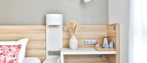 Una habitación de hotel moderna con cabecera de madera, una mesita de noche con lámpara, un florero con flores secas, una cafetera y un teléfono en la mesita de noche.