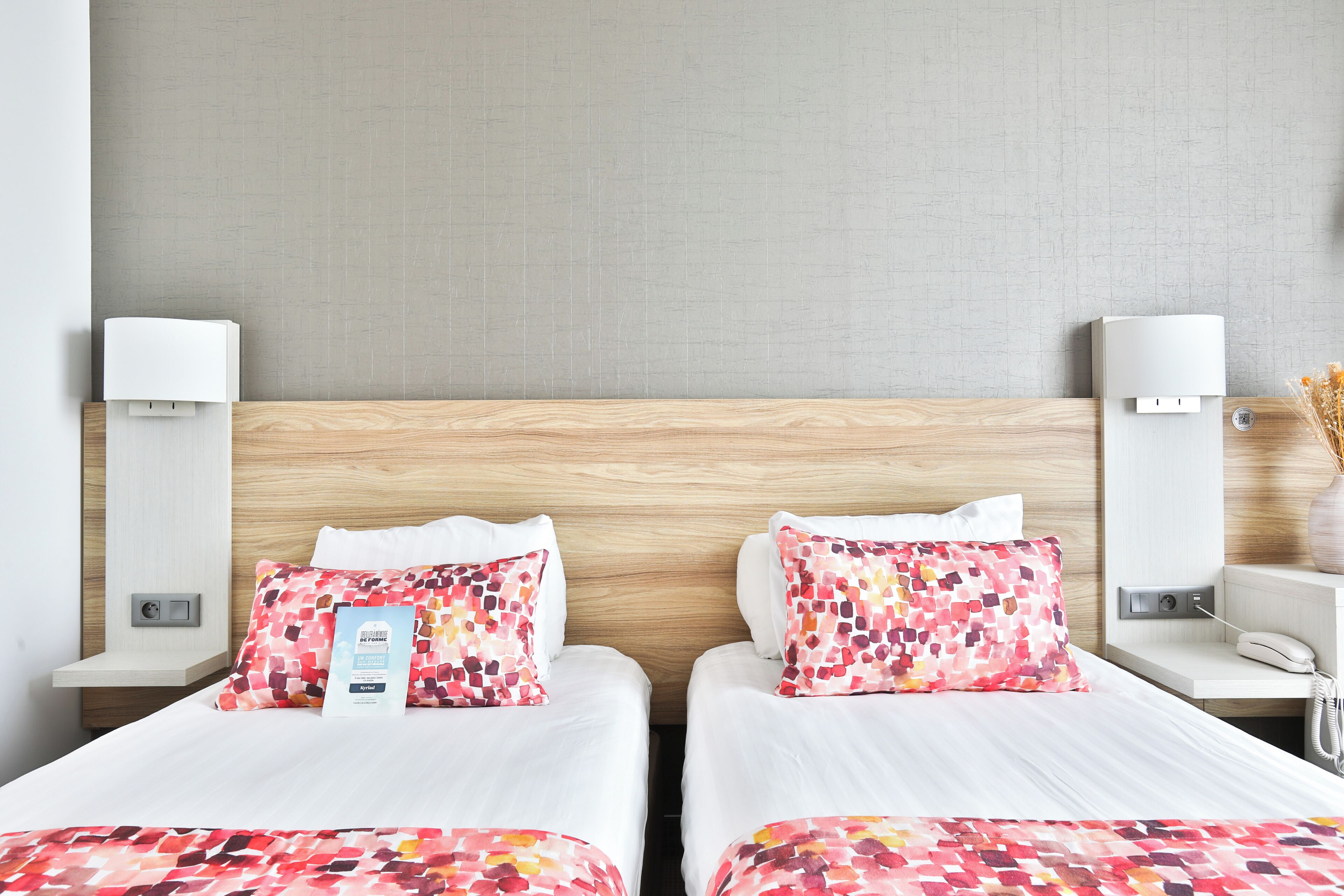 Una habitación de hotel con dos camas, cada una con una colcha blanca y un cojín colorido. Hay mesitas de noche con lámparas y un teléfono.