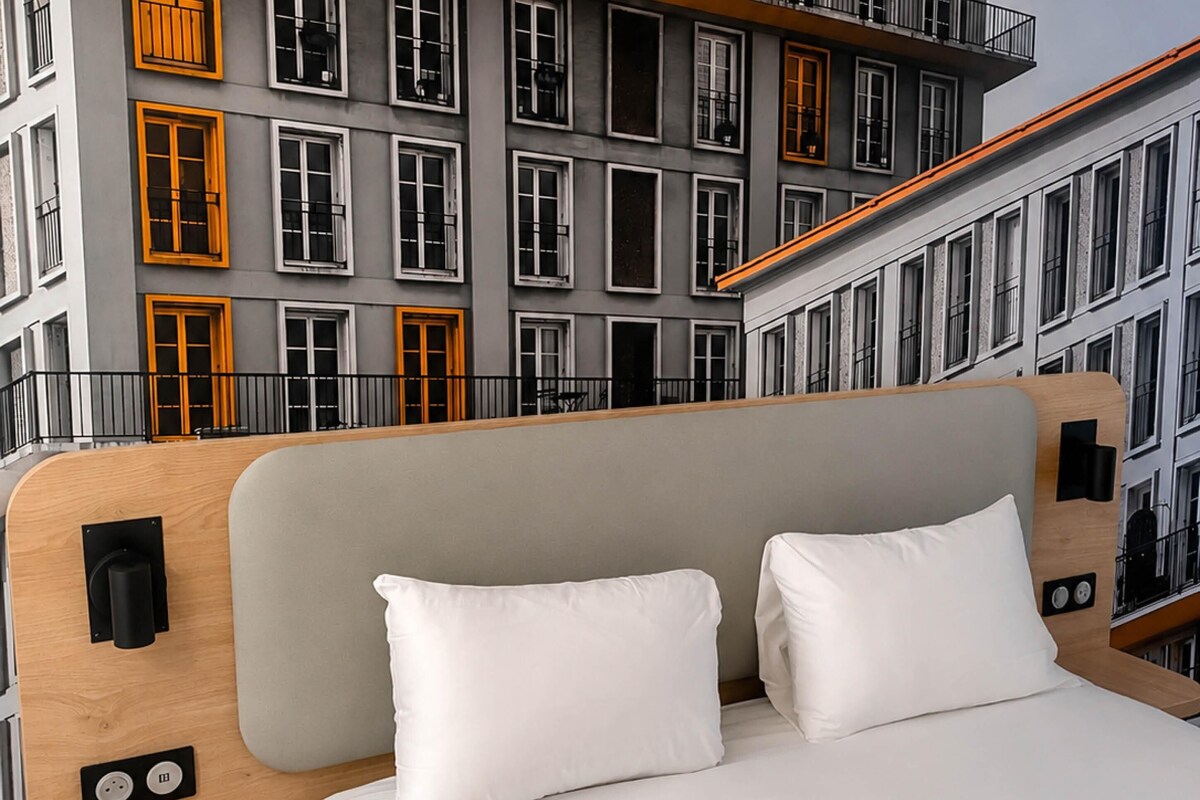 A hotel room with a bed, a wooden headboard, and a view of a building with multiple windows and balconies.