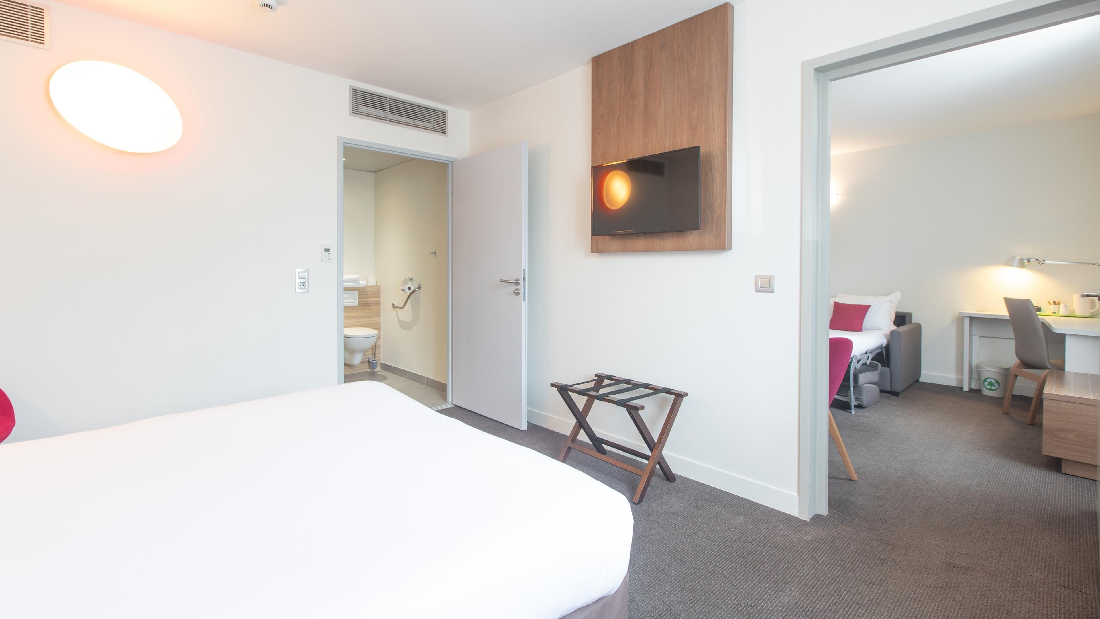 Habitación de hotel con cama, televisor montado en la pared, escritorio con silla y baño visible a través de una puerta abierta.