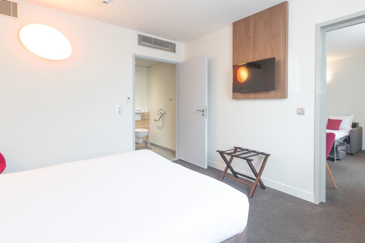 Habitación de hotel con cama, televisor montado en la pared, escritorio con silla y baño visible a través de una puerta abierta.