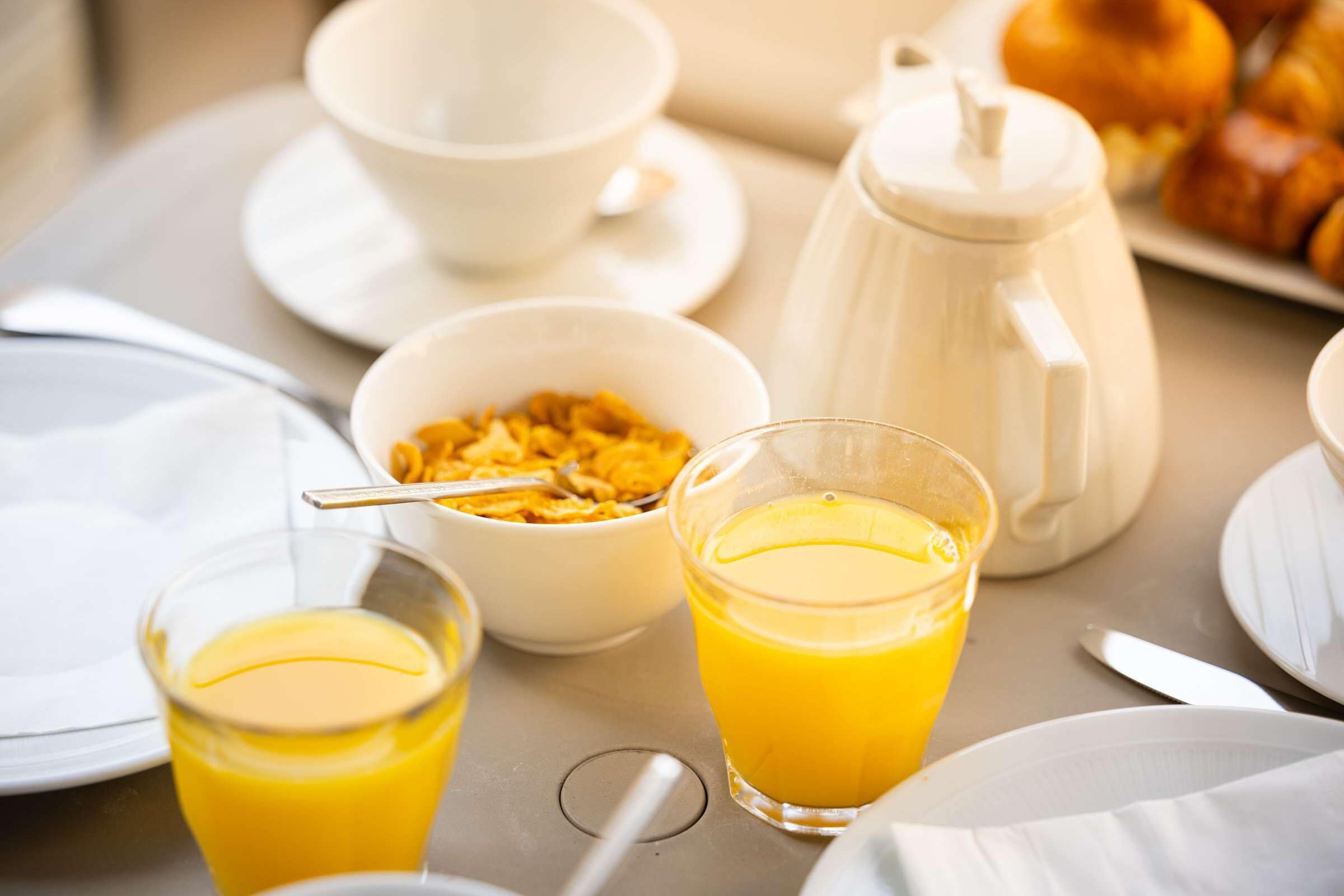 A breakfast setting with a coffee pot, bowls of cereal and orange juice, and croissants on a table.