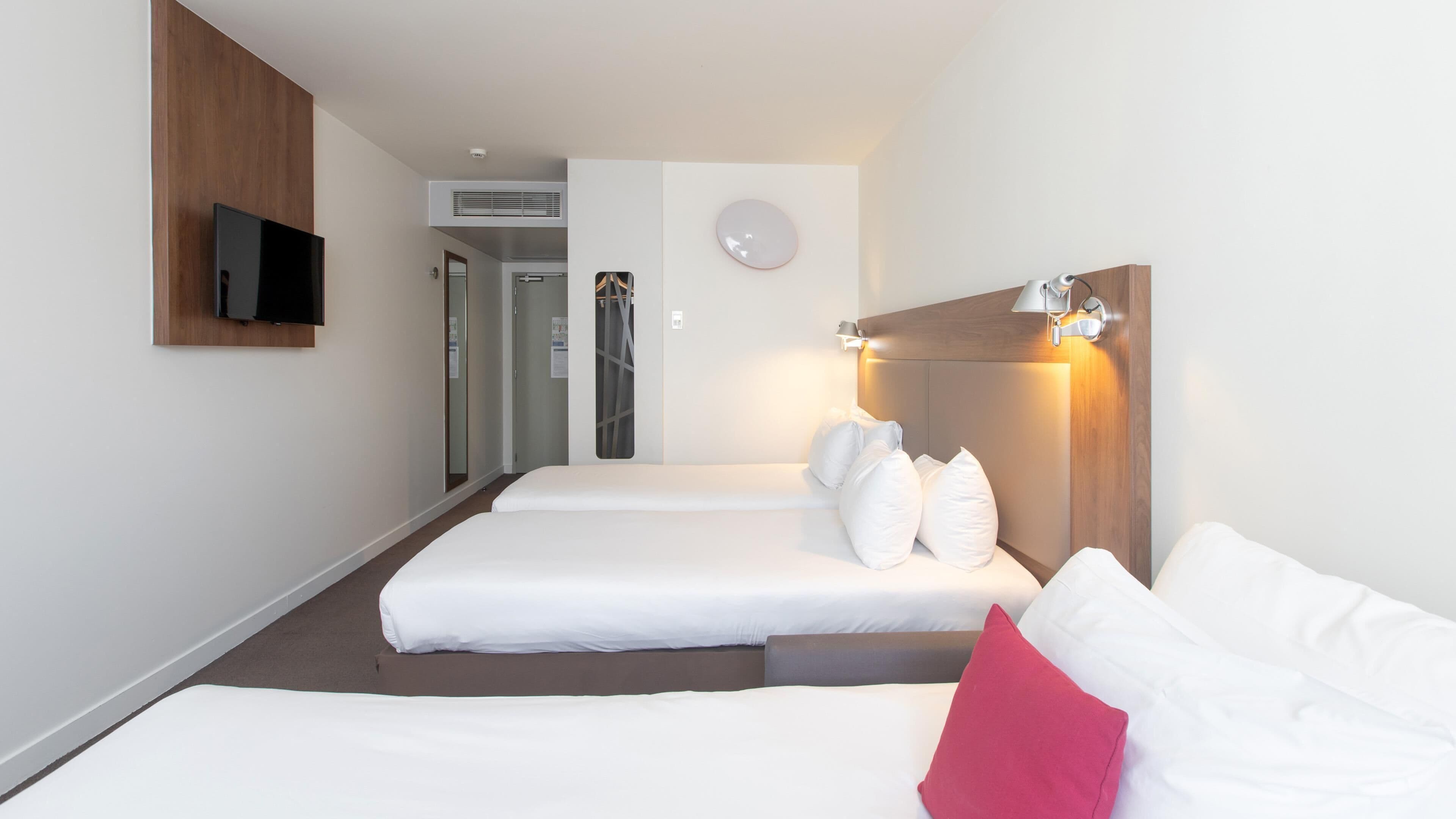 A hotel room with two beds, a television mounted on the wall, a clock, and a wall-mounted light fixture.