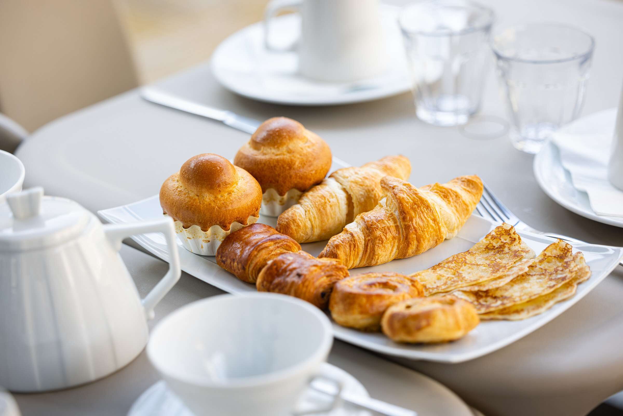 Desayuno con cruasanes, una tetera, tazas y un plato de pasteles sobre una mesa.