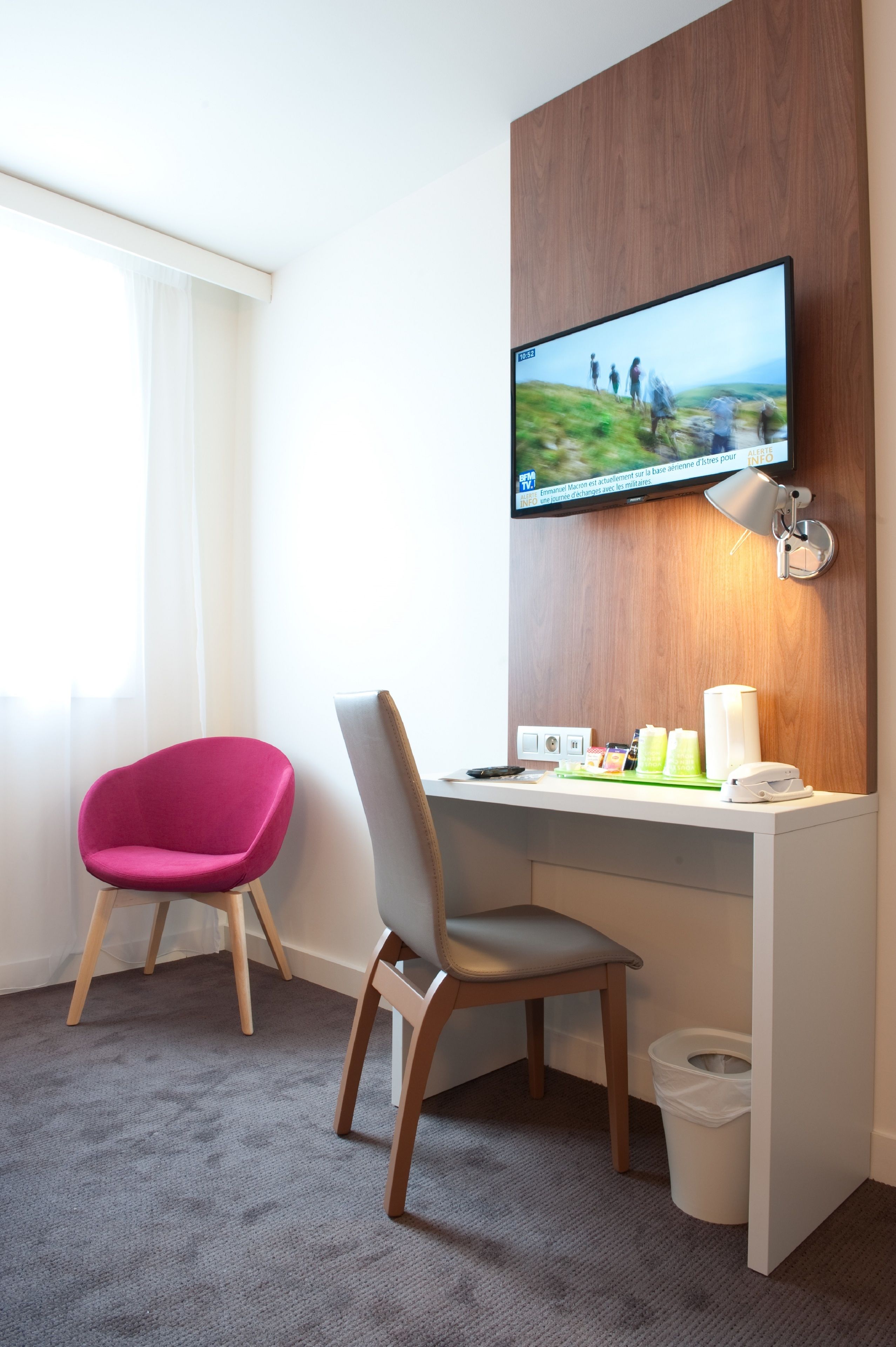 A hotel room with a desk, two chairs, a television mounted on the wall, a waste bin, and a window with curtains.
