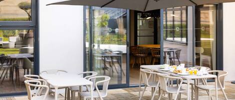 An outdoor dining area with tables and chairs, large umbrellas, and a view through glass doors into a garden.