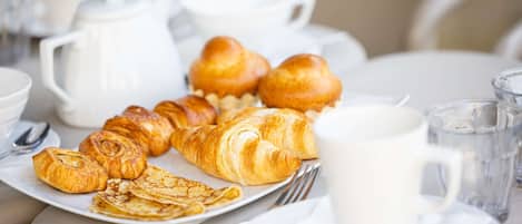 Desayuno con cruasanes, una cafetera, tazas y un vaso de agua sobre una mesa.