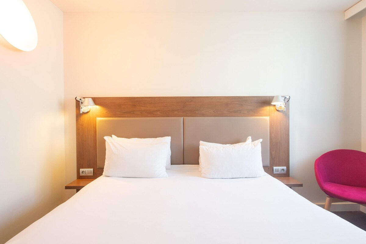 Una habitación de hotel con una cama grande, dos mesitas de noche, un cabecero de madera y una lámpara de pared luminosa.