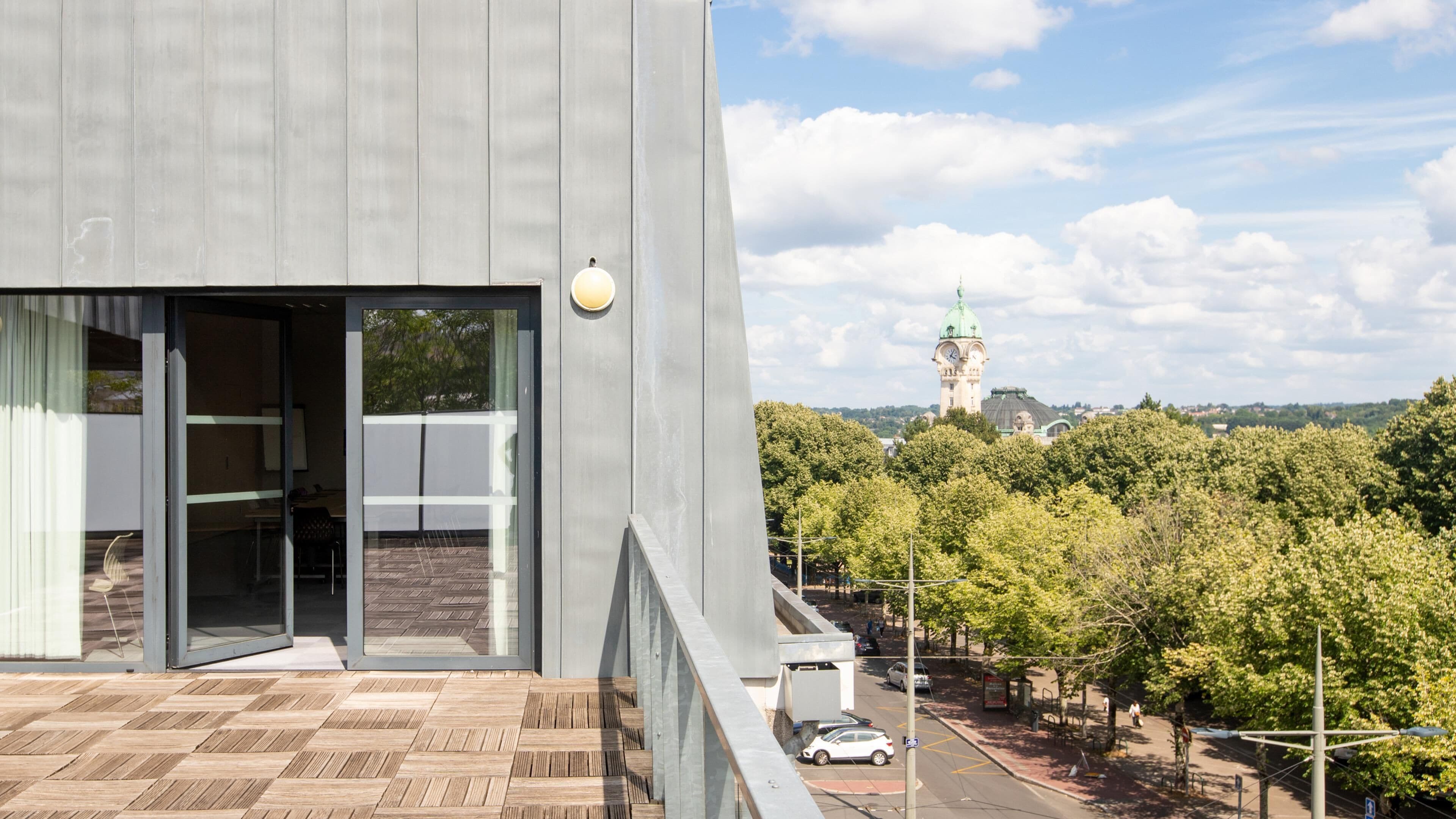 A modern building with a large glass entrance, a balcony with a patterned floor, and a view of a clock tower in the distance.