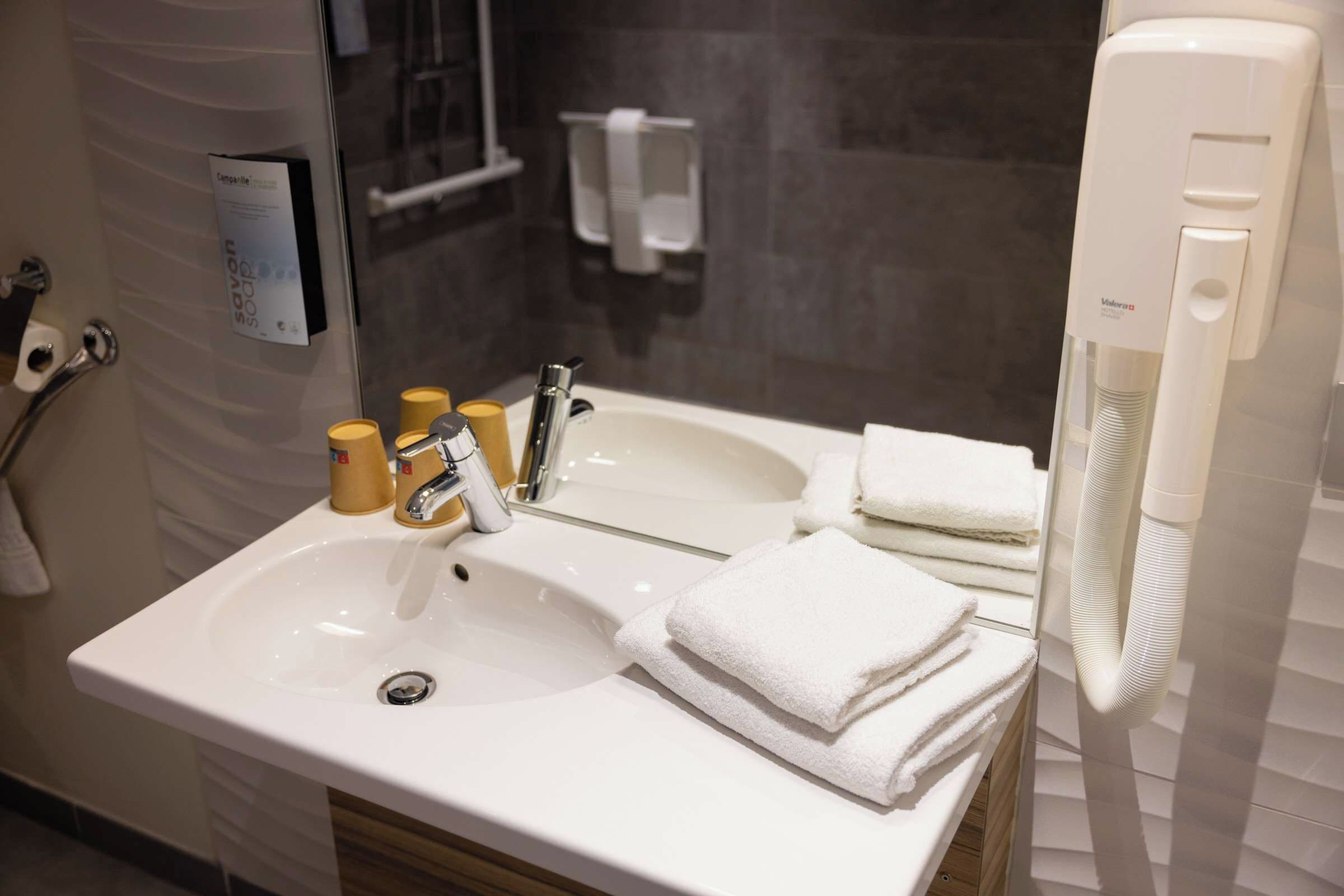 Un bany amb un lavabo blanc, dues aixetes i un escurapulides amb tovalloles doblegades.