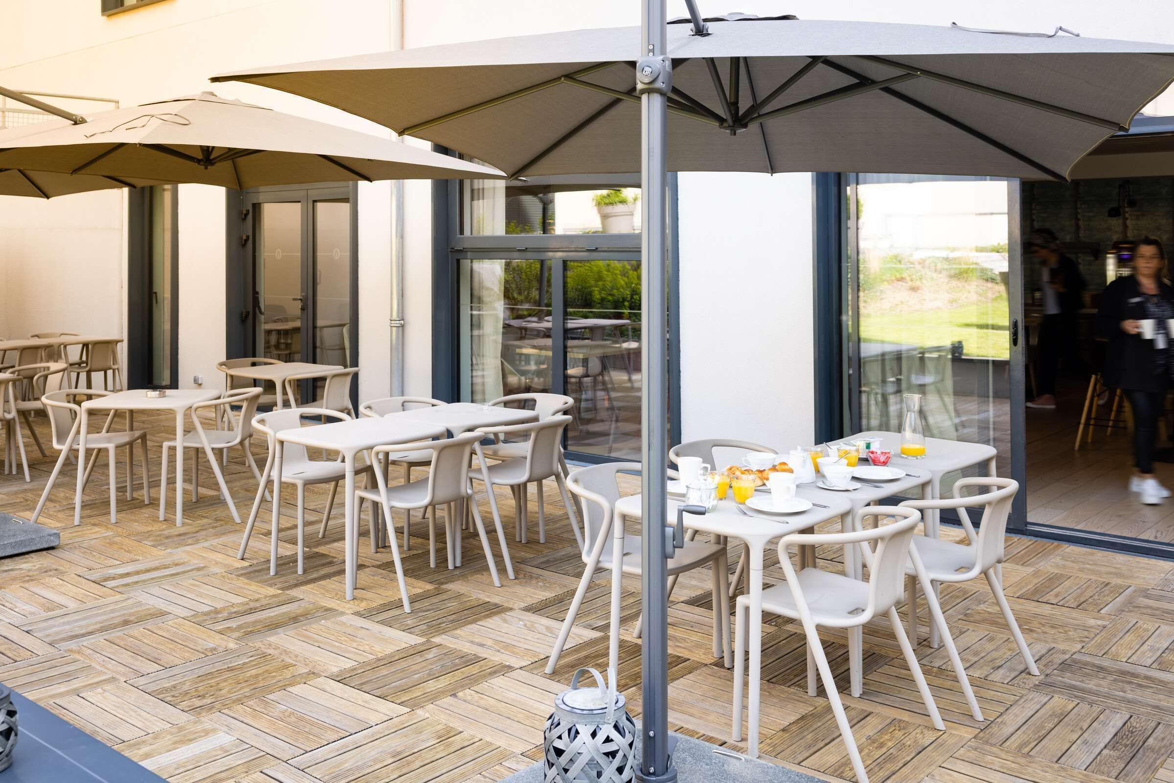 Un espace extérieur aménagé en terrasse, avec des tables et des chaises, un grand parasol et une porte vitrée donnant sur l’intérieur.