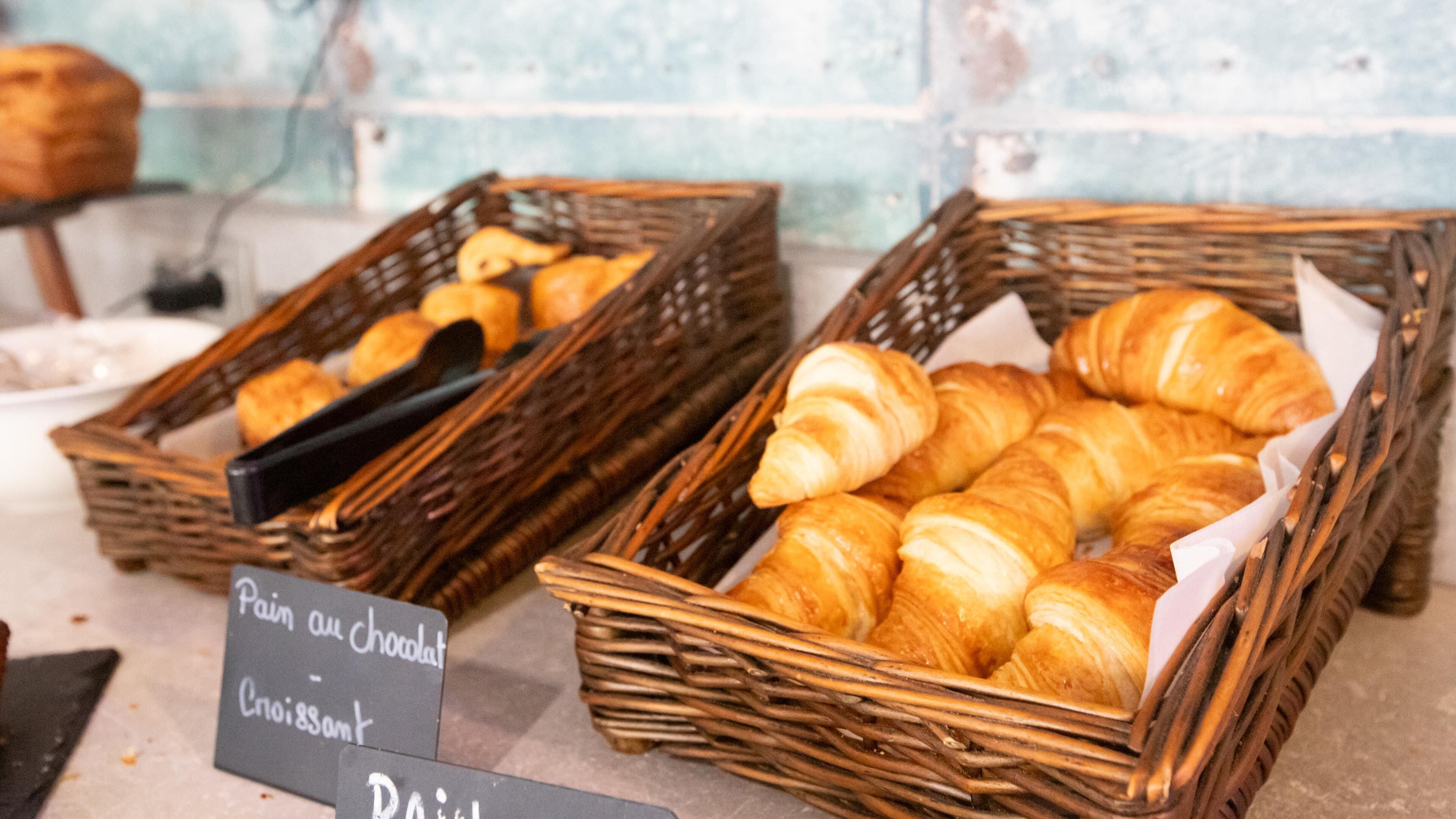 Zwei Korbflechtekörbe, gefüllt mit Backwaren, darunter Croissants, auf einer Theke mit Schildern für 'Pain au chocolat' und 'Croissant'.