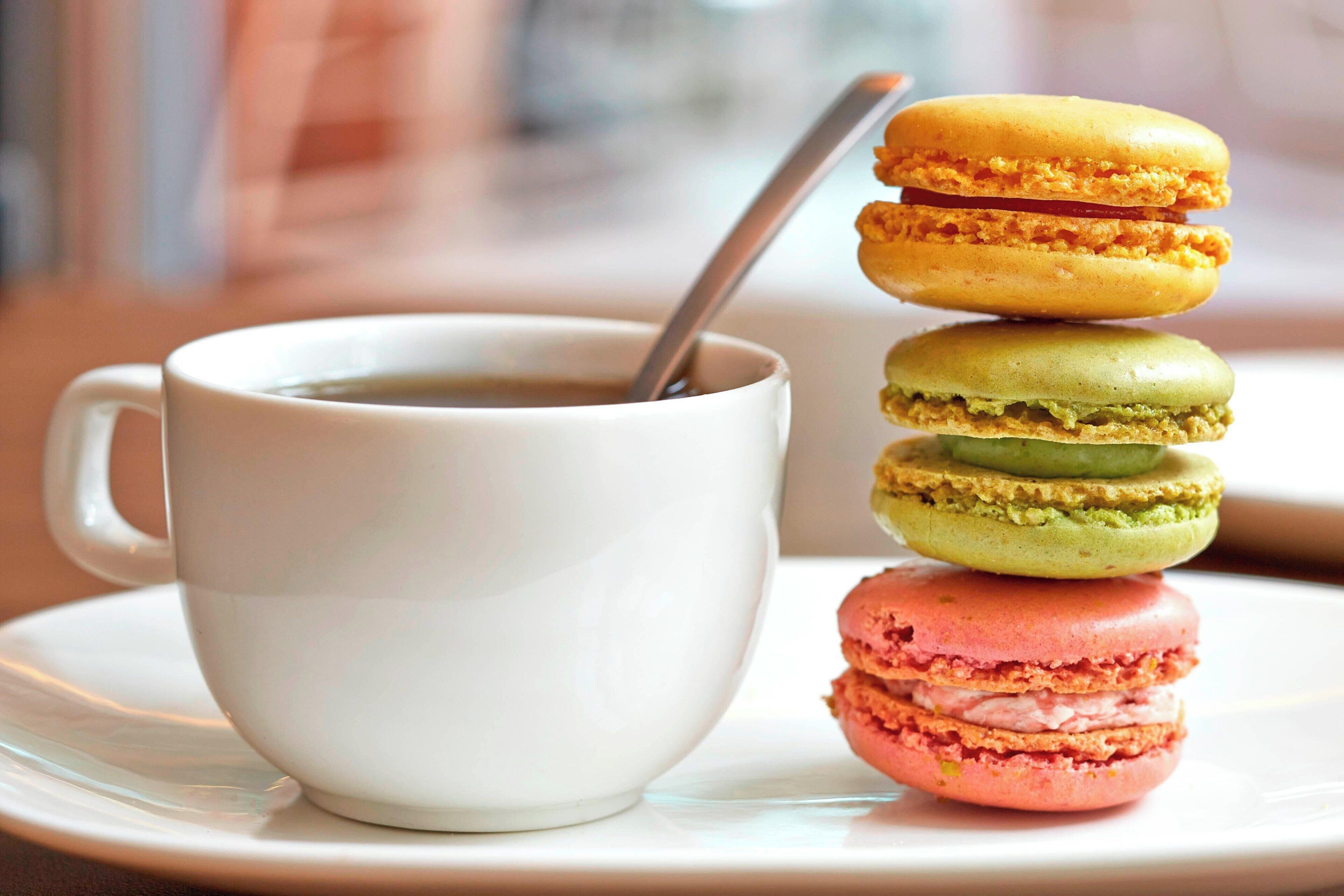 Valkoinen kuppi, jossa on lusikka, päällekkäin aseteltuja värikkäitä macaron-leivoksia ja valkoinen aluslautanen.