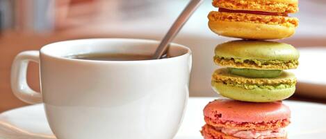Valkoinen kuppi, jossa on lusikka, päällekkäin aseteltuja värikkäitä macaron-leivoksia ja valkoinen aluslautanen.
