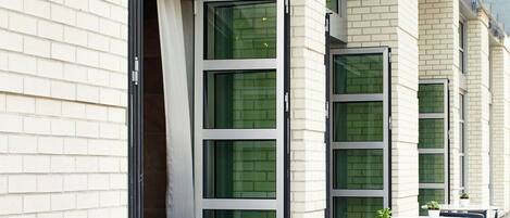 Una hilera de mesas y sillas al aire libre con plantas en macetas, frente a un edificio con ventanas grandes y fachada de ladrillo.