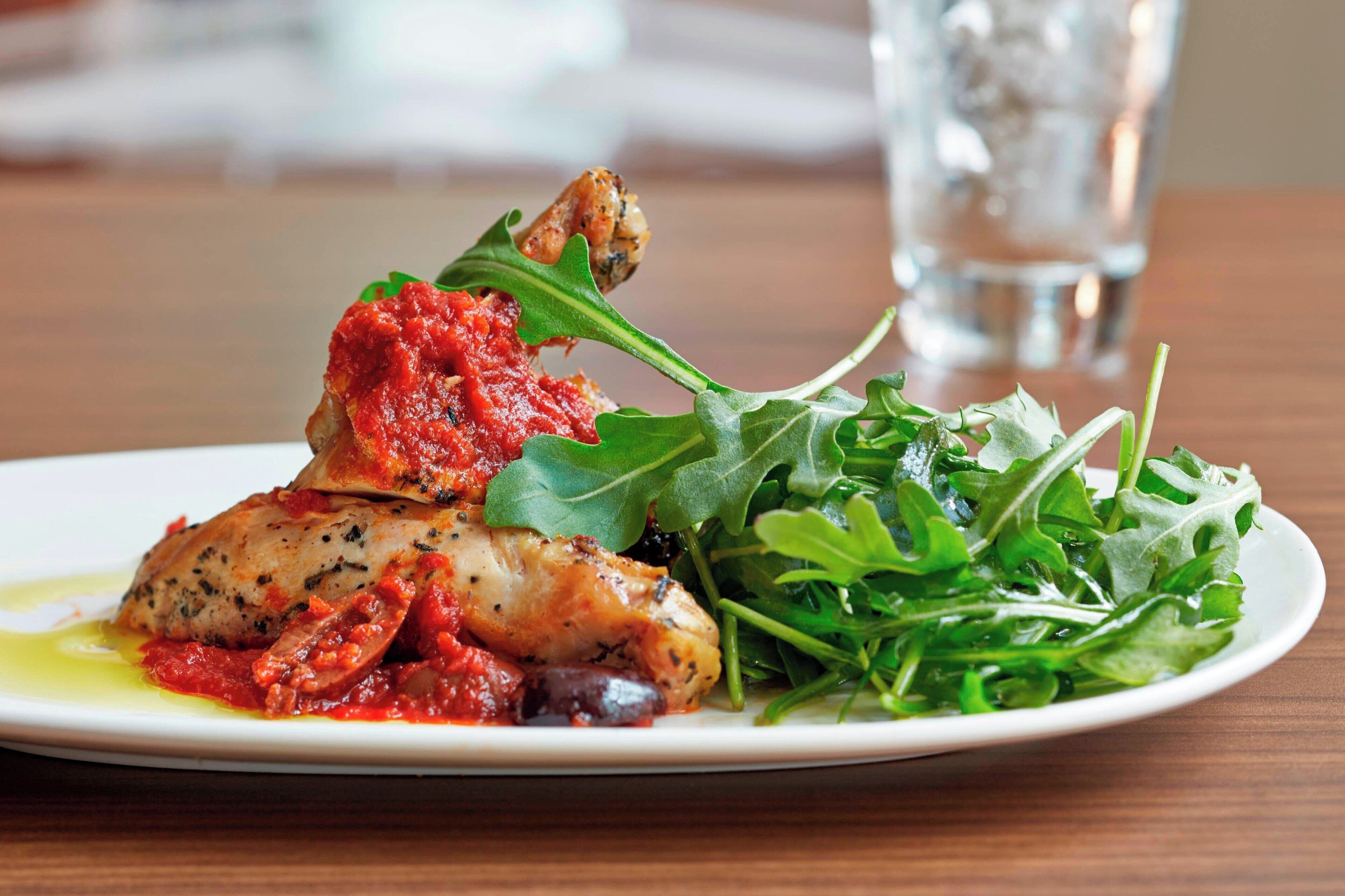 A plate with a piece of chicken, a red sauce, and a green salad on a white plate.
