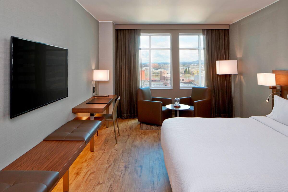 A hotel room with a large bed, a flat-screen TV mounted on the wall, a desk with a chair, a small table with a lamp, and a view of the city through large windows.