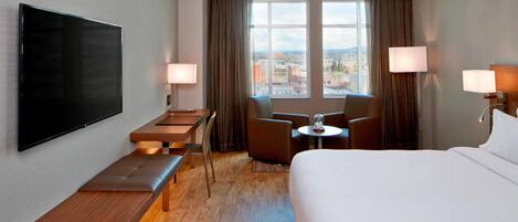 A hotel room with a large bed, a flat-screen TV mounted on the wall, a desk with a chair, a small table with a lamp, and a view of the city through large windows.