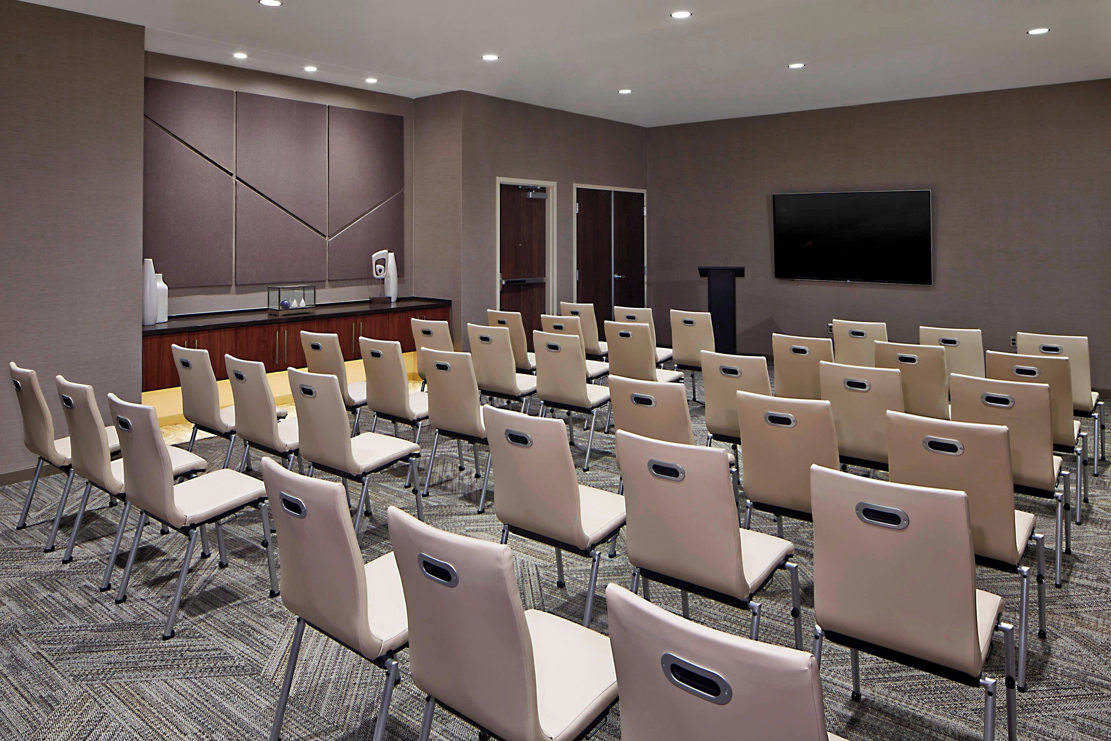 Une salle de conférence avec des rangées de chaises beiges, un pupitre et un téléviseur à écran plat fixé au mur.
