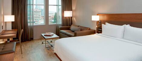 A hotel room with a large bed, a desk with a chair, a small table, and a sofa.