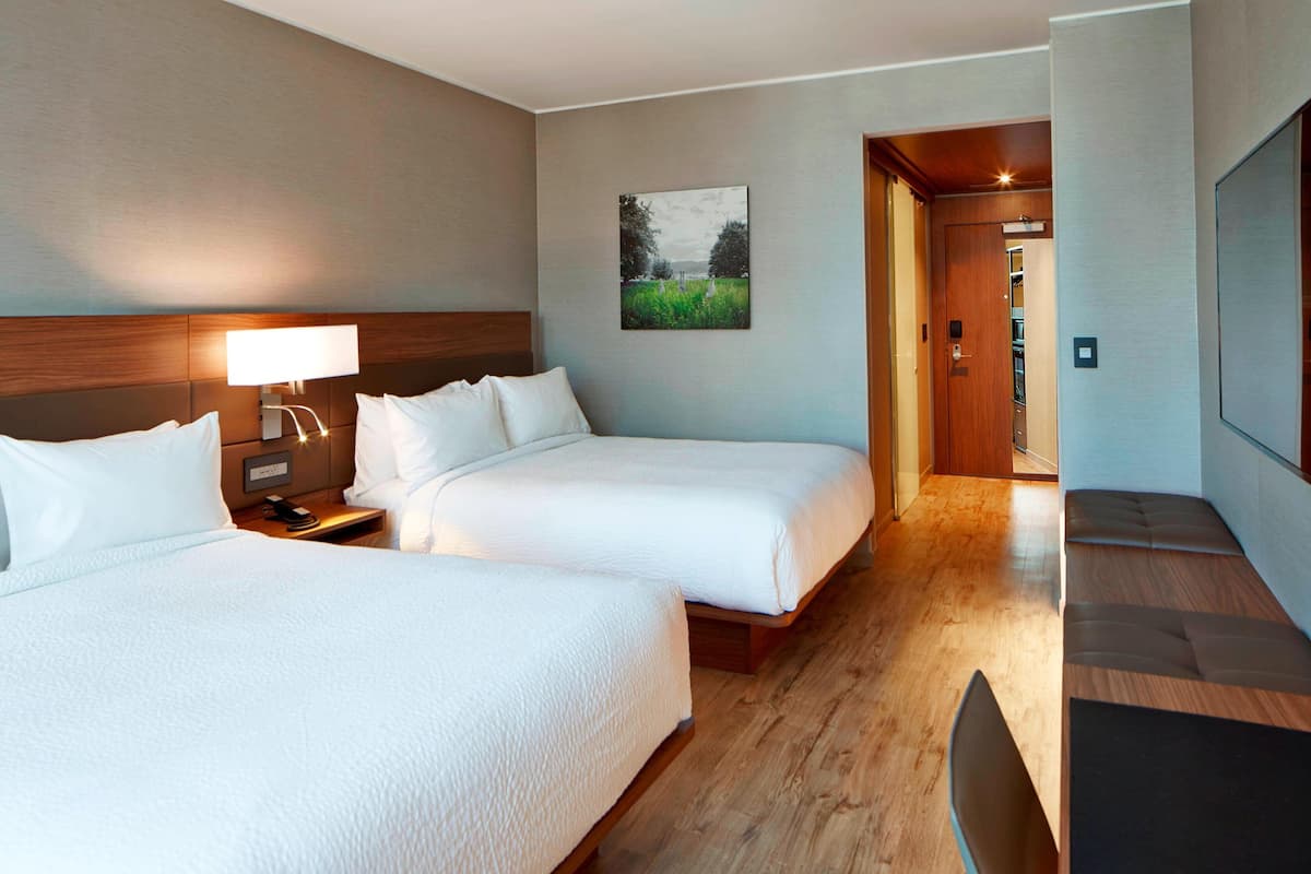 A hotel room with two beds, a nightstand, a lamp, a painting, and a wooden bench.