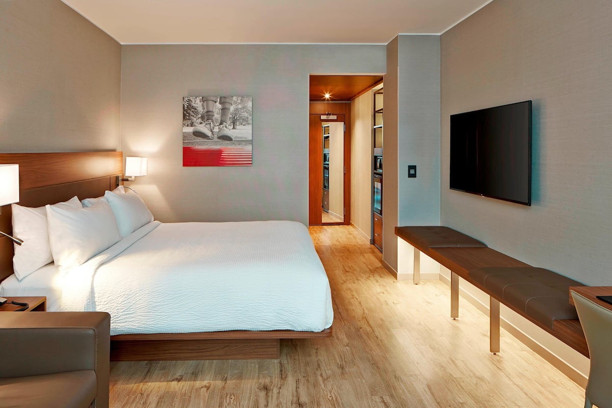 A hotel room with a bed, bedside table, bench, TV, and a hallway leading to a bathroom.